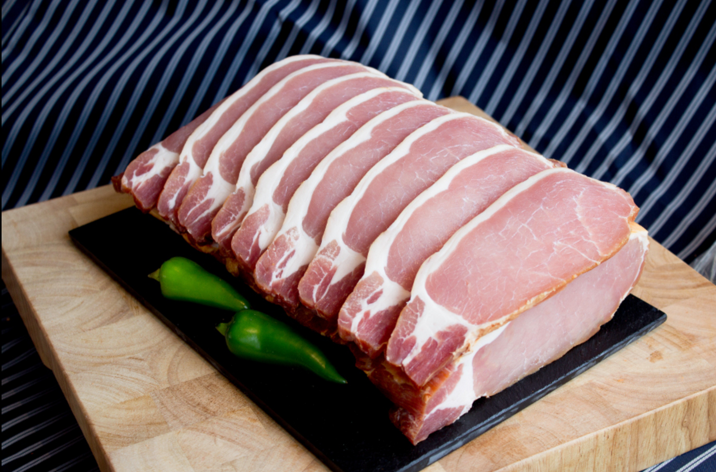 Raw bacon strips on a black slate with green peppers on a wooden board against a striped background
